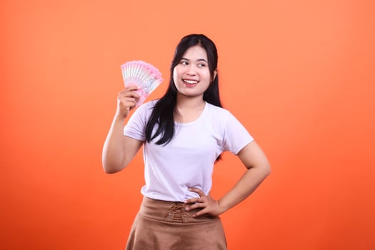 A woman holding a fan of pink paper money isolated on an orange background smiling slightly looking away photo
