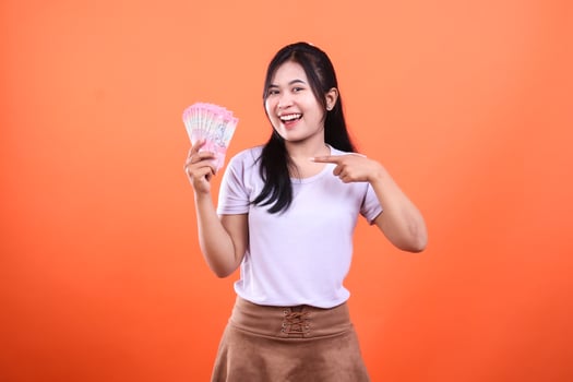 A young woman points to a pile of money, standing in front of an orange backdrop photo