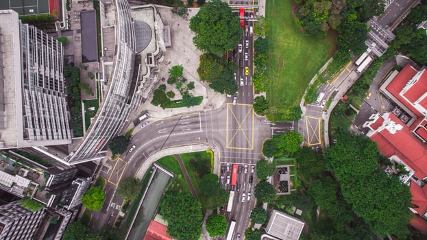 A bustling Singapore intersection with modern buildings, lush greenery, moving vehicles, and a red roofed traditional structure in the foreground. photo