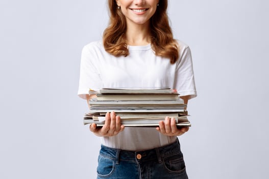 Portrait Of Young Smiling Woman Holding Stack Of Old Magazines In Hands, Millennial Lady Enjoying Sorting Garbage For Recycling, Standing Over Light Background In Studio, Cropped Shot With Free Space photo