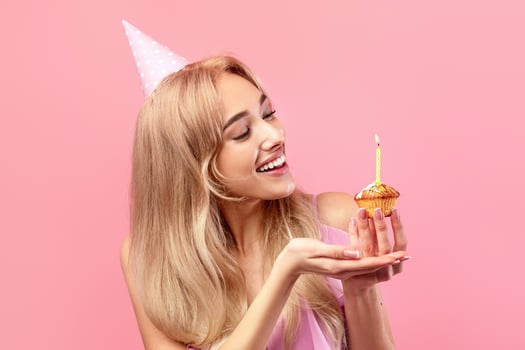 Beautiful millennial woman in birthday cap eating tasty cake, celebrating B-day on pink studio background. Blonde lady enjoying yummy dessert, having fun on holiday. Festive concept photo