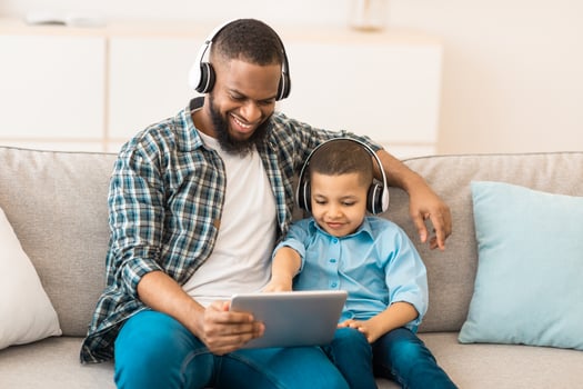 African American Dad And His Son Using Digital Tablet Wearing Headphones Watching Cartoons Online Sitting On Sofa At Home. Family Bonding Using Computer Together On Weekend photo