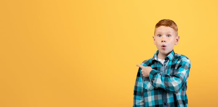 Amazed red-haired school boy in plaid shirt aiming at copy space over yellow studio background. Shocked ginger kid pointing at empty space, showing exciting advert for children photo