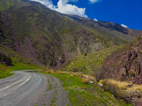Winding gravel road meanders through rocky Kyrgyzstan mountains adorned with various colors of rock and vegetation. The serene landscape evokes feelings of peace and adventure under a bright sunny sky. photo