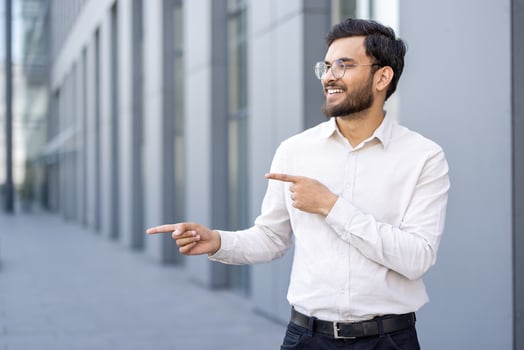 A smiling businessman of Indian descent points sideways, wearing glasses and a button-down shirt, outdoors with a modern building in the background. photo