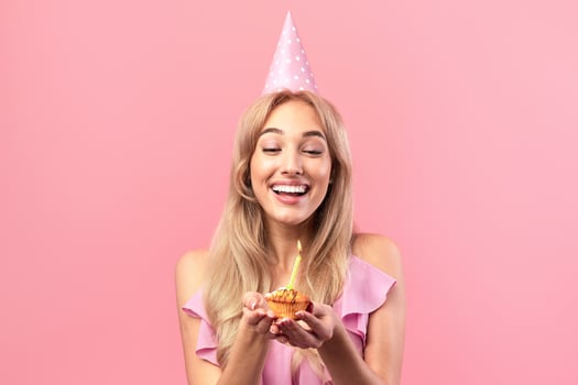 Portrait of joyful blonde woman in cute dress celebrating and holding birthday muffin with candle on pink studio background. Happy millennial lady with heart shaped balloon eating festive dessert photo
