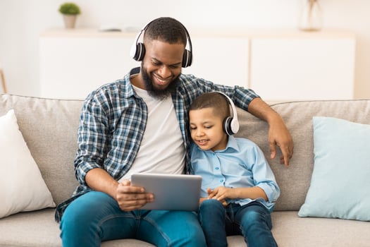 African American Father And Son Using Digital Tablet Wearing Wireless Headphones Watching Cartoons Online Together Sitting On Couch At Home. Family Weekend Leisure, Gadgets And Modern Lifestyle photo
