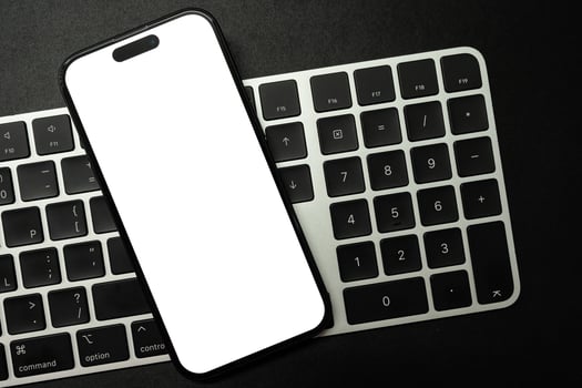 A modern smartphone with a blank white screen rests on a white and black computer keyboard, ideal for showcasing digital content or technology concepts. Copy space. photo