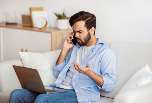 Computer Problems. Frustrated arab freelancer guy talking on cellphone and looking on laptop screen at home, calling customer support hotline, suffering bad internet connection while working online photo