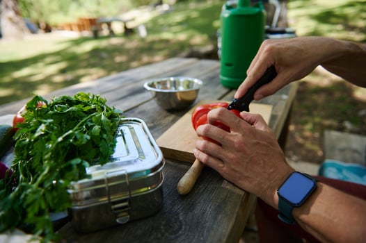 Person Preparing Vegetables Outdoors on a Wooden Table with Fresh Herbs and Utensils photo