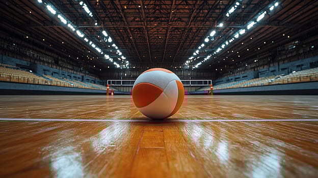 A lone volleyball rests on the gleaming wooden court of an empty stadium, awaiting the start of a match. The overhead lights cast a dramatic glow across the grandstand seating. photo