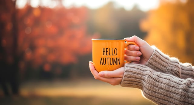 Hands hold an orange mug with Hello Autumn text amidst fall foliage. Cozy season, warm drinks, and nature. photo