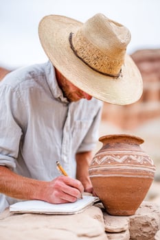 Male archaeologist wearing a straw hat is documenting findings in a notebook beside a traditional clay pot, showcasing the process of cultural exploration and preservation photo