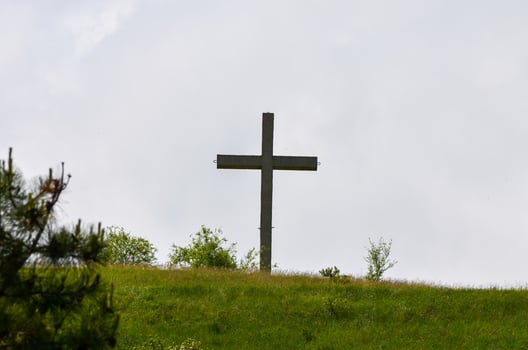 A wooden cross is standing on a hillside in a grassy field. The sky is cloudy, and the cross is the only visible object in the scene photo