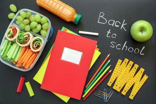 School supplies and lunch box with tasty rolls, cucumbers, carrots, grapes, apple, bottle of juice on black background, back to school concept. Top view. photo