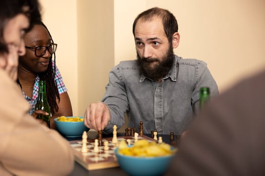 Selective focus on caucasian man holding chess piece, concentrating on his female opponent during their game. Bearded white guy sits at table with snacks and chessboard, planning his next move. photo