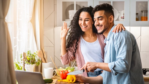 Middle Eastern Food Bloggers Couple Filming Content In Kitchen For Their Culinary Vlog, Happy Arab Man And Woman Cooking Healthy Lunch Together And Waving Hands And At Laptop Camera, Greeting Viewers photo