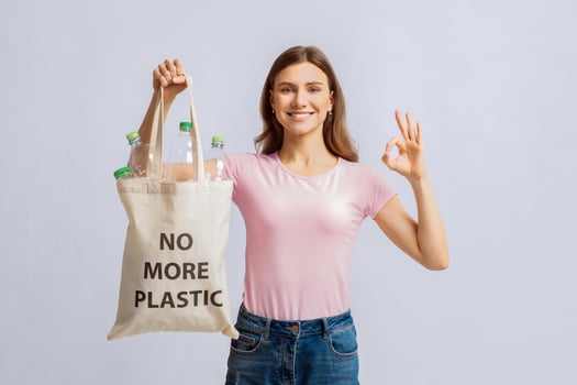 Portrait Of Positive Young Lady Holding Eco Bag With No More Plastic Inscription And Empty Bottles Inside, Showing Ok Gesture, Cheerful Millennial Woman Posing On Light Studio Background, Free Space photo