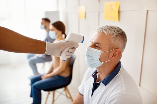 Senior man in medical mask waiting for covid-19 vaccination at hospital, doctor checking his temperature with infrared thermometer. Immunization against coronavirus, population protection photo