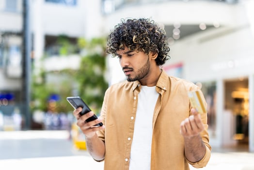 A man looking concerned while holding a credit card and smartphone in a shopping mall setting. photo