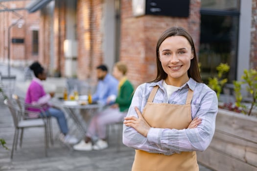A smiling waitress in an apron poses in front of her outdoor restaurant, customers sit at tables in the background. photo