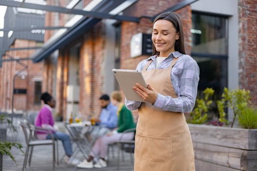 A smiling waitress takes an order at an outdoor restaurant with a tablet, overseeing customers. photo