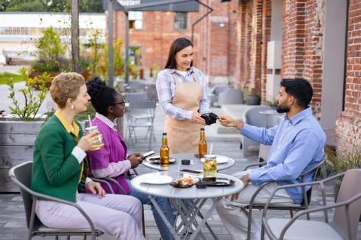 A female waiter hands a portable terminal to a customer at a restaurant. People are sitting at a table. photo