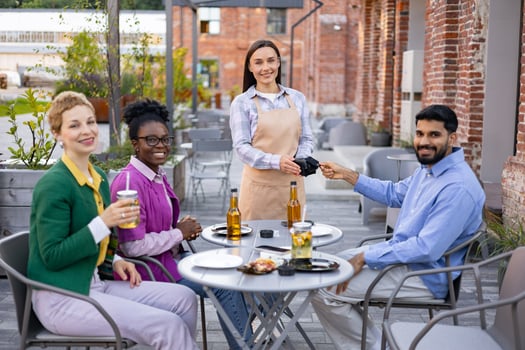 A female waiter in an apron serves customers at an outdoor restaurant, accepting payment via a portable terminal. photo
