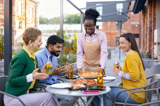 A diverse group of friends enjoying pizza and drinks at an outdoor restaurant, served by a friendly waitress. photo