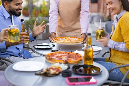 A diverse group of people enjoying pizza, drinks, and each other's company outdoors. A waiter is serving pizza. photo