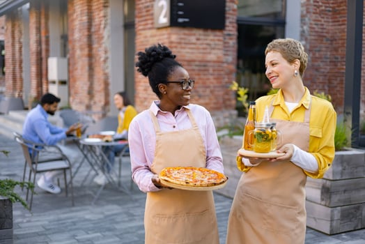 Two waitresses smiling, carrying pizza and drinks outdoors at a restaurant, with customers in the background. photo