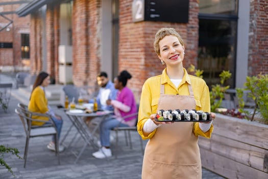 A smiling waitress presents sushi rolls outdoors on a patio. Customers dine in the background. photo
