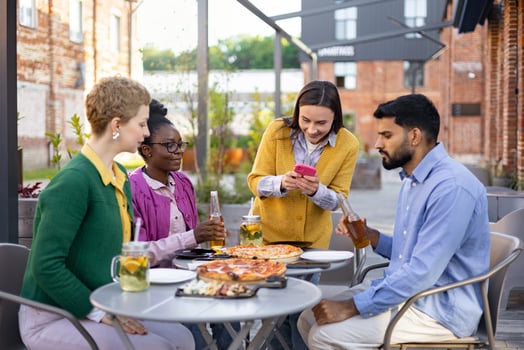 Diverse group of friends enjoying pizza and drinks together at an outdoor restaurant, smiling and using a phone. photo