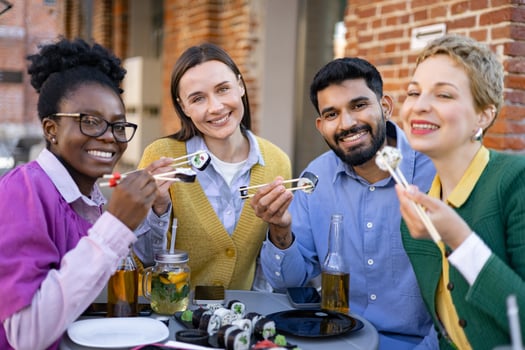 A diverse group of friends enjoys sushi together, smiling and laughing while eating outdoors on a sunny day. photo