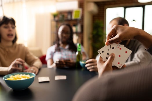 African american hand displaying winning card combo while others focus on game. Young adults seated at table with beer bottles and snacks, having relaxed bonding time, playing games. photo