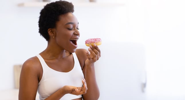 Fit Black Lady Having Cheat Meal Eating Donut Enjoying Unhealthy Tasty Sweet Food Standing In Kitchen At Home. Weight Gain Issue, Sweets Overeating, Bad Nutrition Habit Concept. Side View photo
