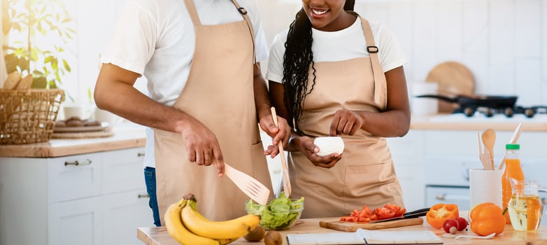 Young Happy African American Lovers In Aprons Cooking Together In Kitchen, Cheerful Black Couple Enjoying Preparing Healthy Food At Home, Adding Seasoning To Fresh Vegetable Salad, Free Space photo