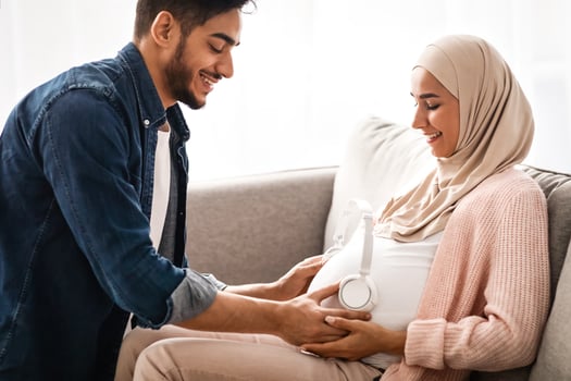 Music For Baby In Womb. Loving Arab Husband Placing Wireless Headphones On Pregnant Wife's Belly While They Resting Together On Couch In Living Room At Home, Muslim Spouses Enjoying Future Parenthood photo