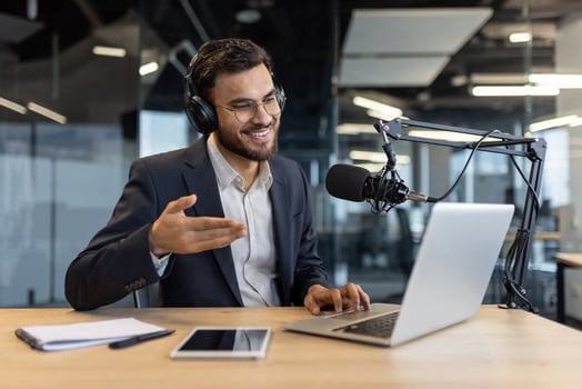 A businessman in a suit smiles while podcasting, wearing headphones, using a laptop, and speaking into a microphone in a modern office setting. photo