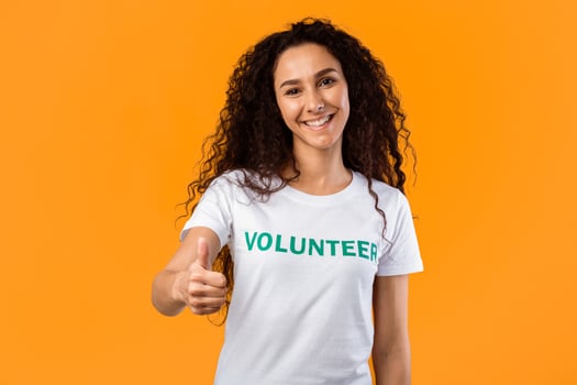 Happy Female Volunteer Gesturing Thumbs Up Approving Charity Work Posing Smiling To Camera Standing In Studio Over Yellow Background. Activist Lady Showing Thumb In Approval. I Like Volunteering. photo