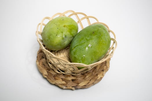 Two fresh green mangoes placed in a woven bamboo basket on a braided mat, set against a clean white background. photo