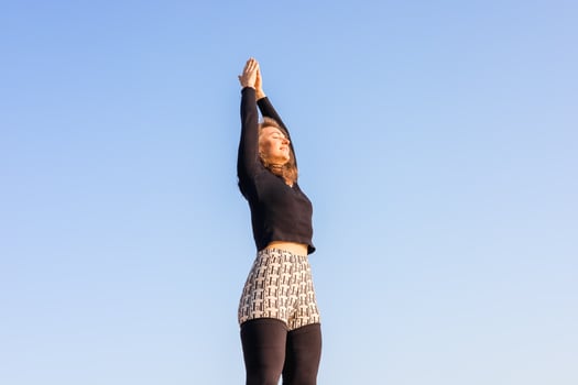 A young Caucasian woman with long brown hair stretches her arms upward against a clear blue sky. She wears a black top and patterned shorts. photo