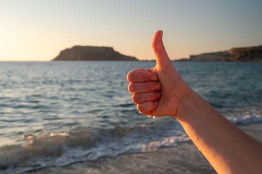 Hand gesturing thumbs up sign on beach at sunset with island in background photo