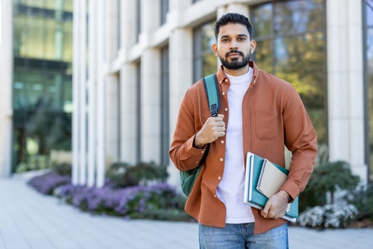 A student stands outdoors, holding books and a backpack in front of a modern building. He is ready for class. photo