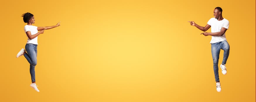 Excited african-american couple jumping up and cheerfully pointing at empty space between them, orange studio background. Emotional young black man and woman aiming at advertisement, panorama photo