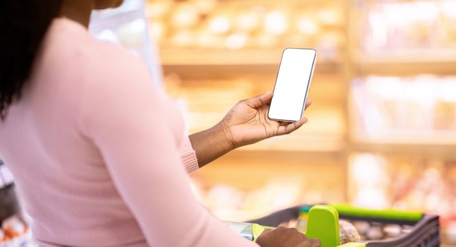 Unrecognizable black woman with shopping cart holding smartphone with blank screen at supermarket, mockup for grocery list app design. Closeup of young lady with mobile device at store photo