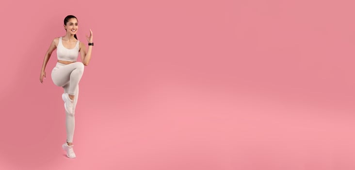 Aerobics Sport Exercises And Healthy Lifestyle. Portrait Of Smiling Slim Young Lady Lifting Leg Up, Doing High Knees Cardio Exercise Standing Isolated On Pink Studio Background, Full Length photo