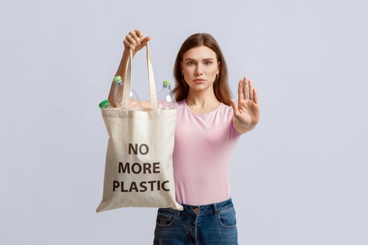 Woman holding eco shopping bag with no more plastic inscription filled with bottles and showing stop gesture with open palm, environmental activist standing over light background in studio photo