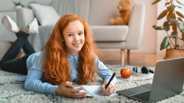 Online Education. Positive red-haired teenage girl lying on the floor carpet with laptop and writing in notebook. Pupil watching lecture or webinar, studying at home using internet, looking at camera photo