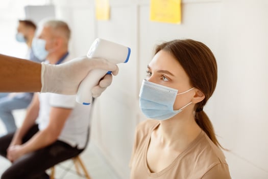 Young woman in face mask waiting for covid-19 immunization at hospital, doctor checking her temperature with infrared thermometer. Vaccination against coronavirus concept photo